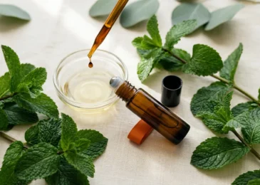 A glass dropper releasing a single drop of peppermint essential oil into a shallow dish of golden carrier oil, beside an open amber roller bottle and fresh peppermint leaves.